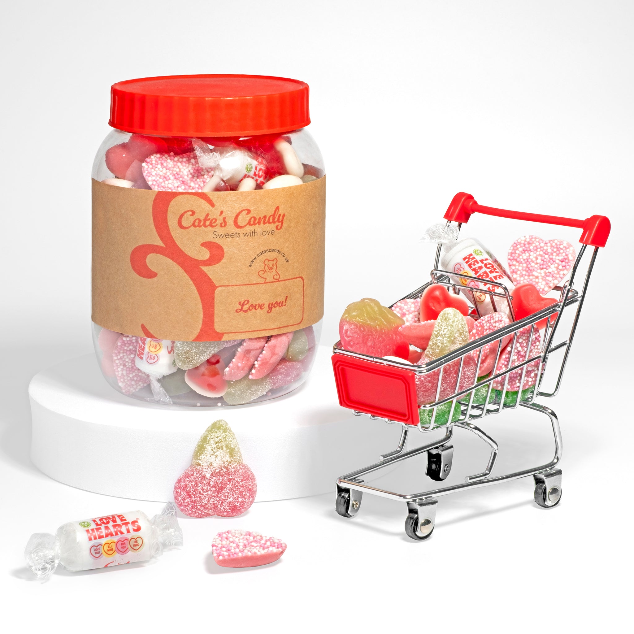  Jar of assorted sweets with a red lid and a small shopping trolley filled with fizzy, jelly and sour sweets on a white background.
